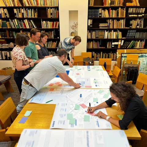 Een groep van zes mensen schrijft samen ideeën uit op grote vellen papier die op een tafel liggen.